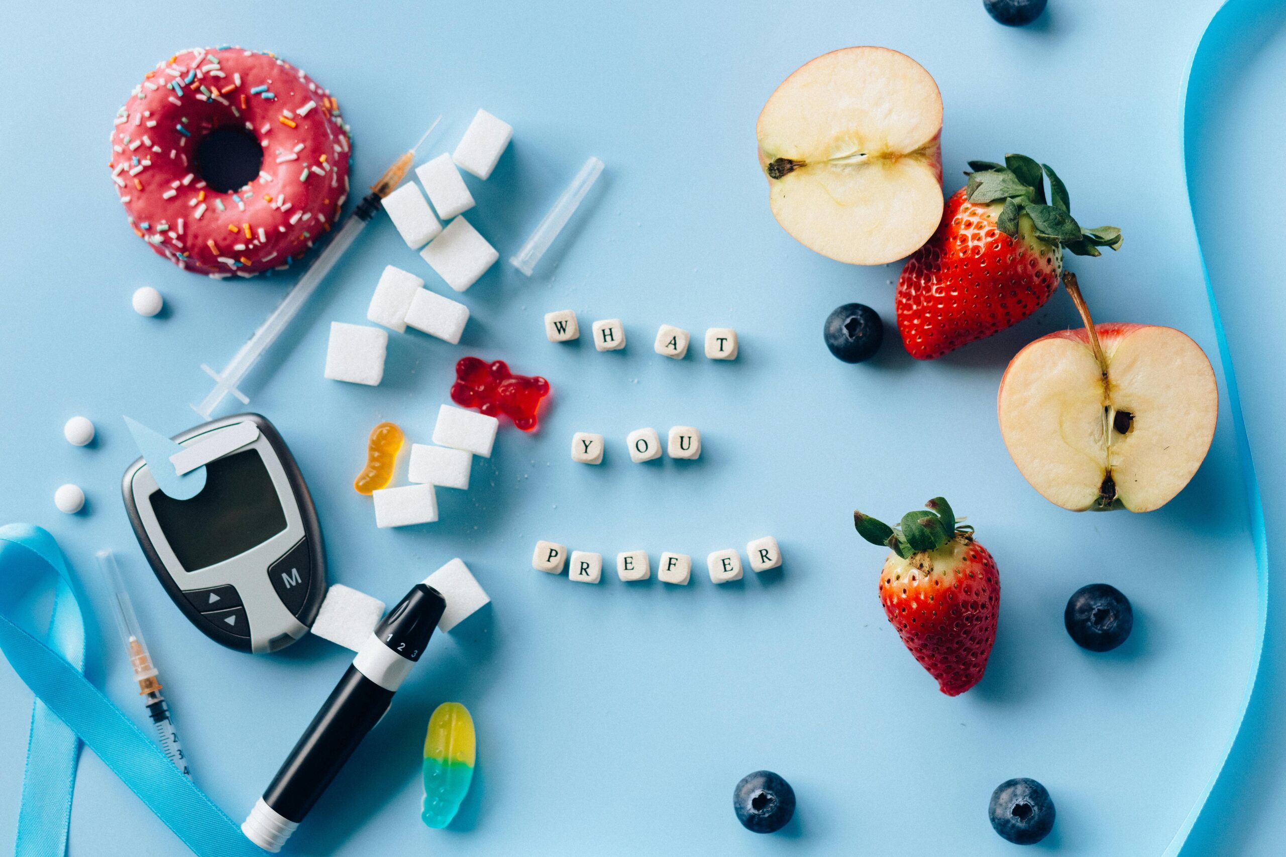 A flat lay comparison of sugary treats and fresh fruits symbolizing healthy choices. The image features a pink donut with sprinkles, sugar cubes, syringes, a glucometer, and gummy candies on one side, contrasting with apples, strawberries, blueberries, and a blue ribbon on the other. The phrase "What You Prefer" is spelled out with letter tiles in the center.